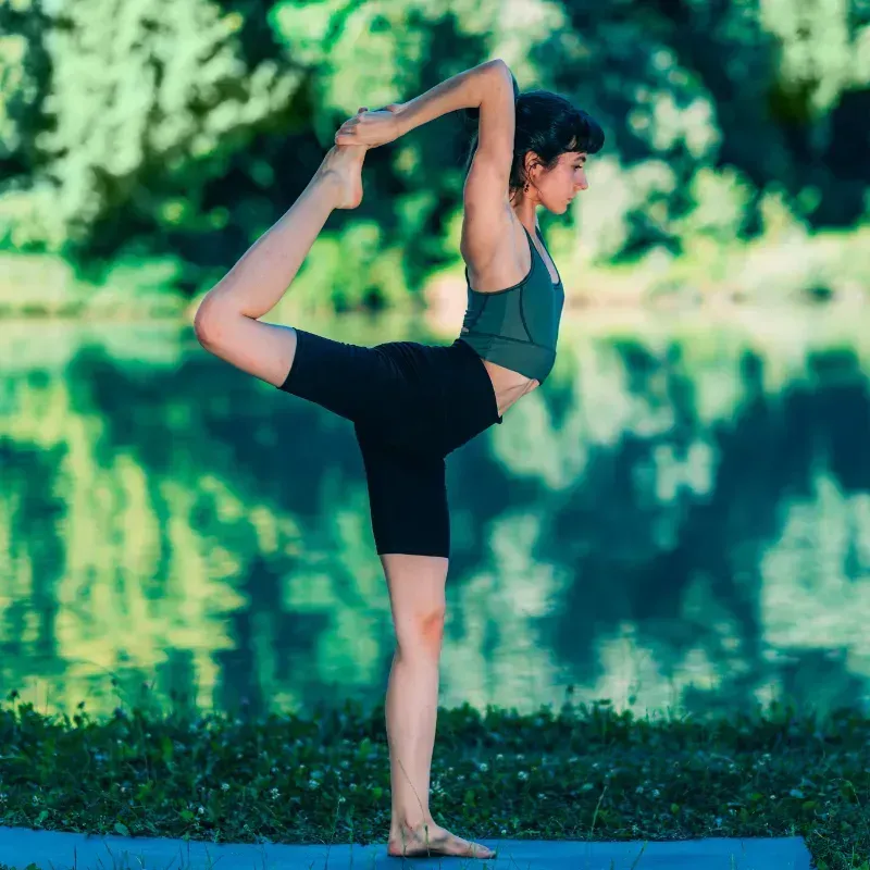 Una mujer está de pie sobre una pierna sobre una colchoneta de yoga frente a un lago.
