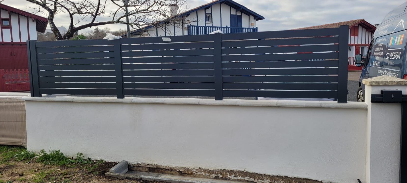 Valla metálica gris sobre un muro blanco, frente a una casa en una calle