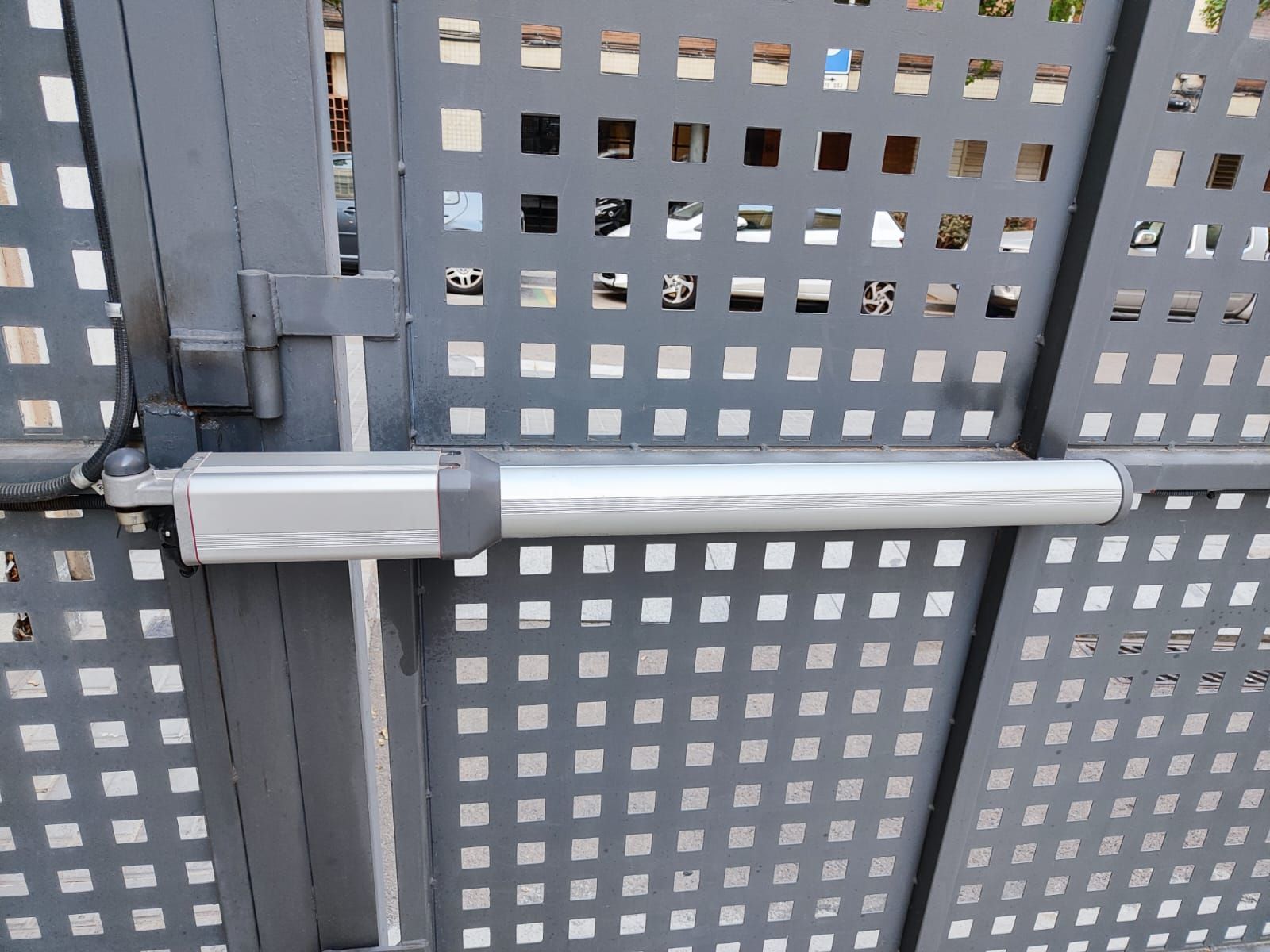 Cierrapuertas horizontal plateado montado en una puerta metálica gris perforada