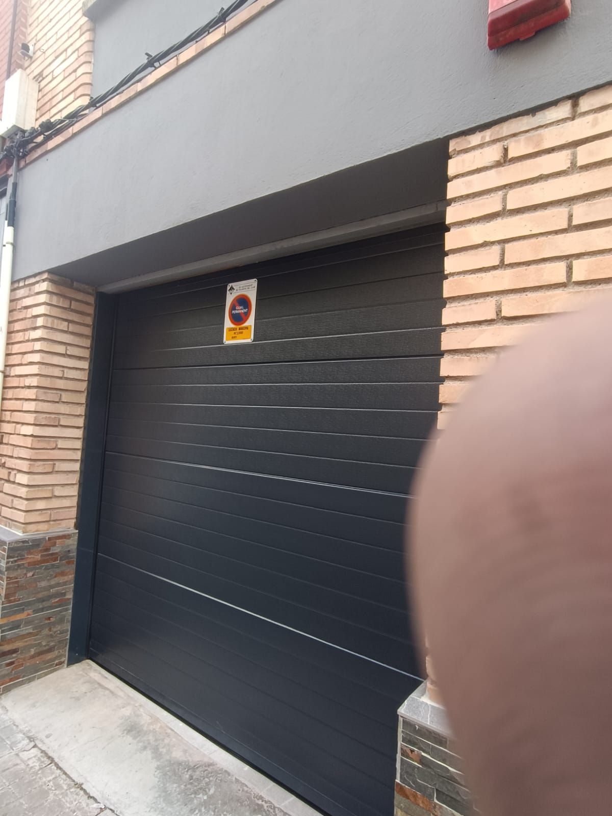 Puerta de garaje negra en una fachada de ladrillo, con una señal de advertencia encima.