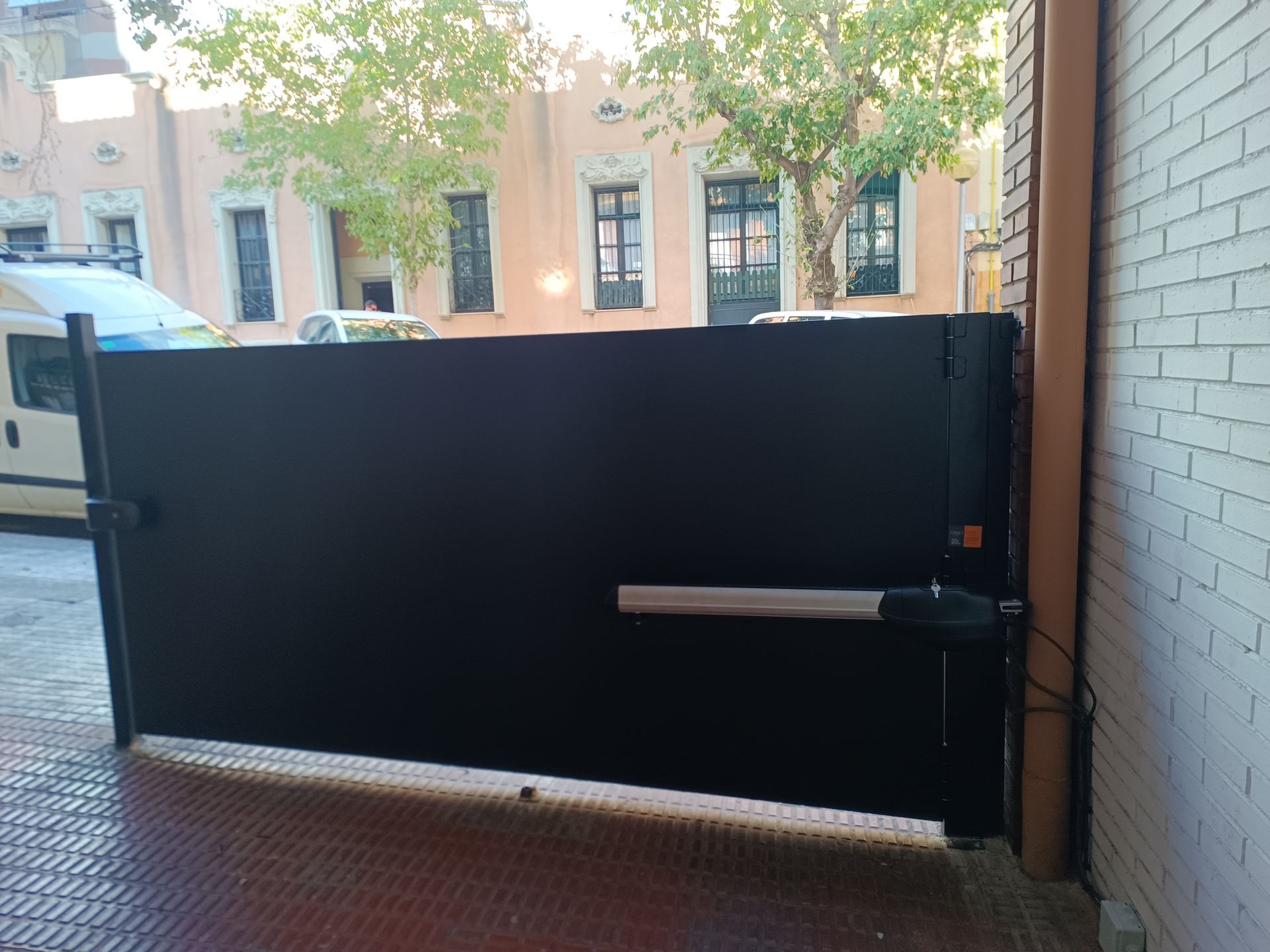 Una verja negra de metal bloquea una entrada de ladrillo, con una furgoneta plateada y edificios visibles al otro lado