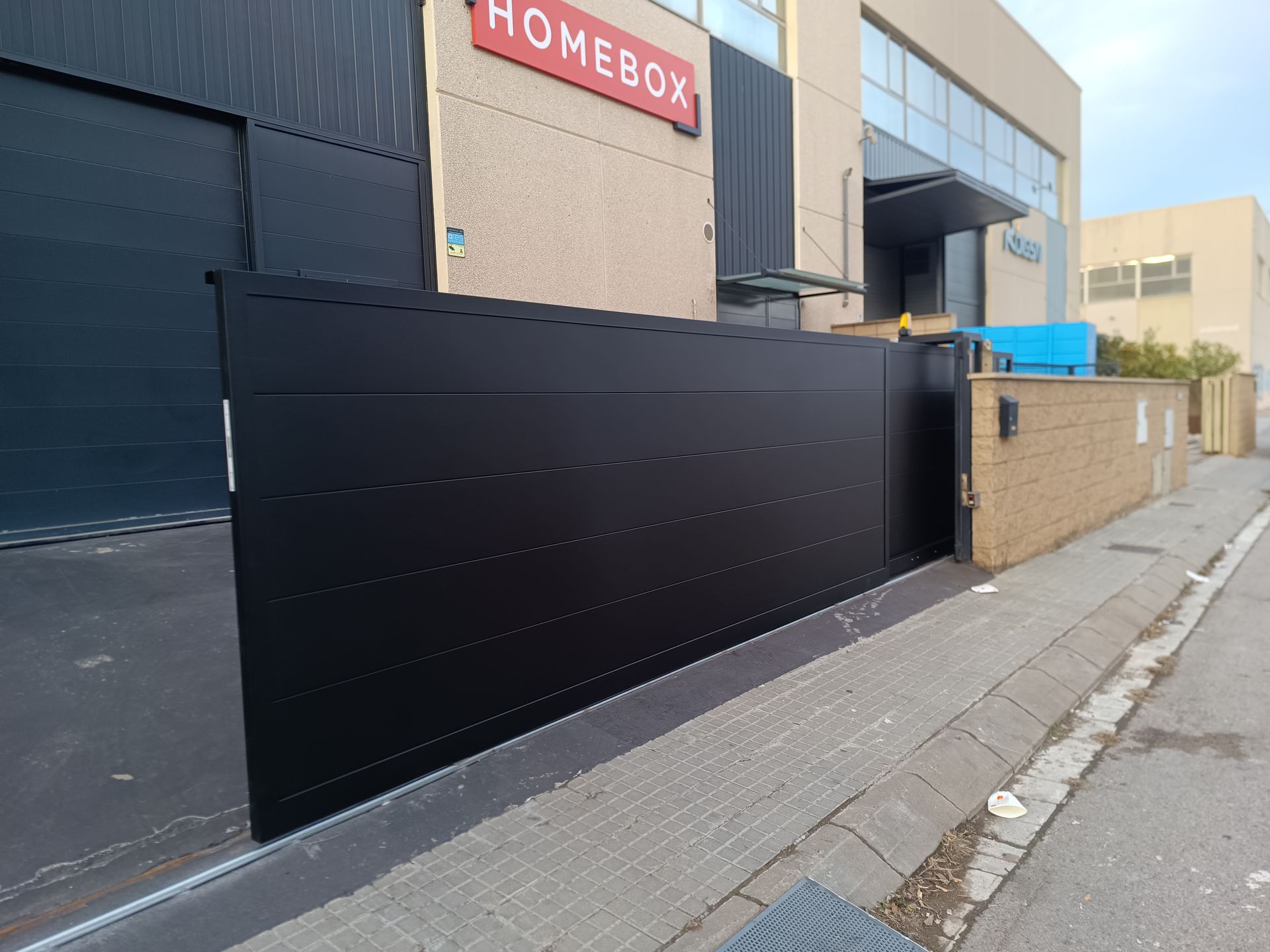 Puerta corredera negra frente a un edificio beige con un letrero rojo de HOMEBOX, junto a una acera y una calle