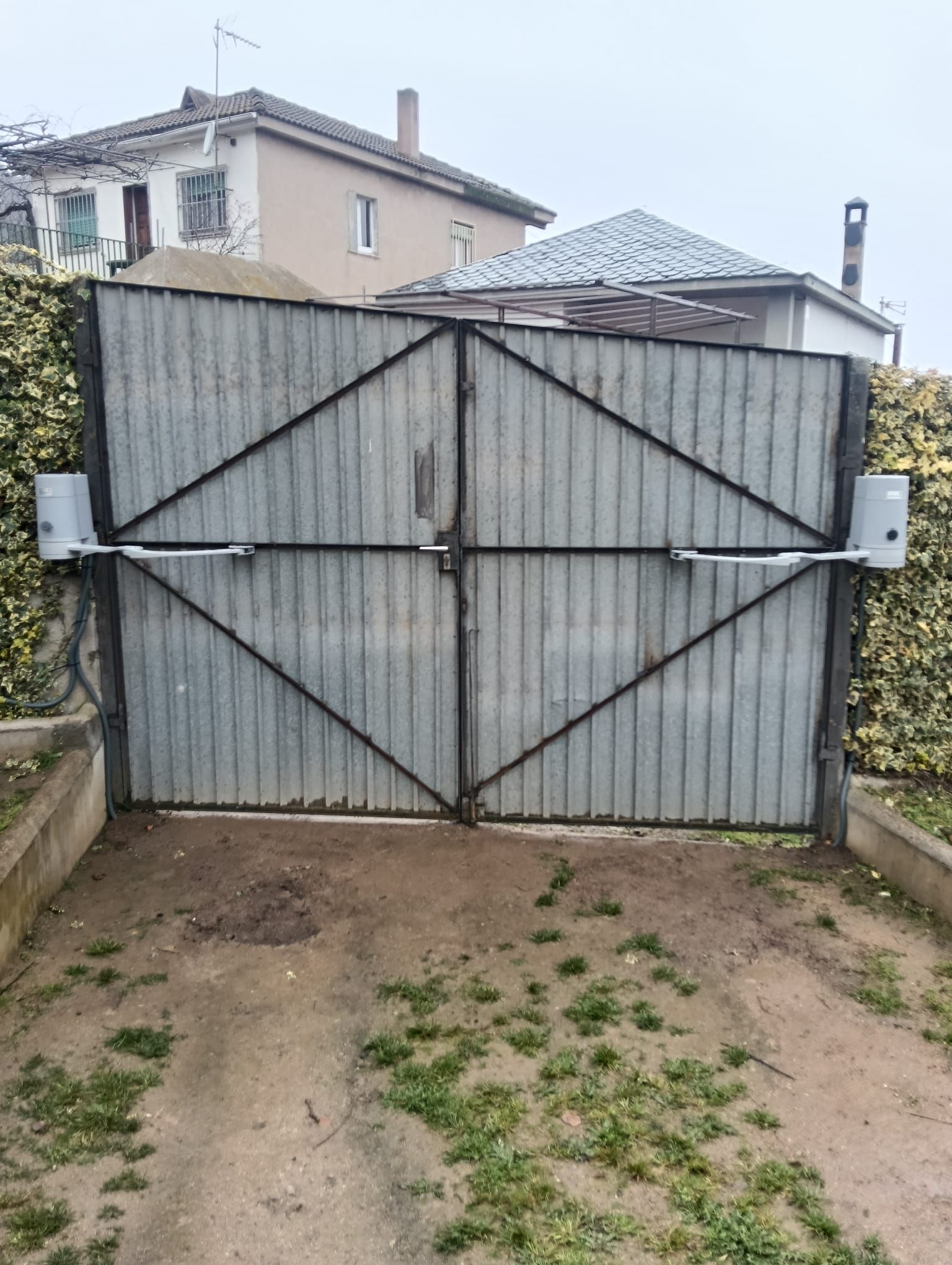 Portón cerrado de metal corrugado gris que bloquea una entrada para vehículos, con casas visibles detrás
