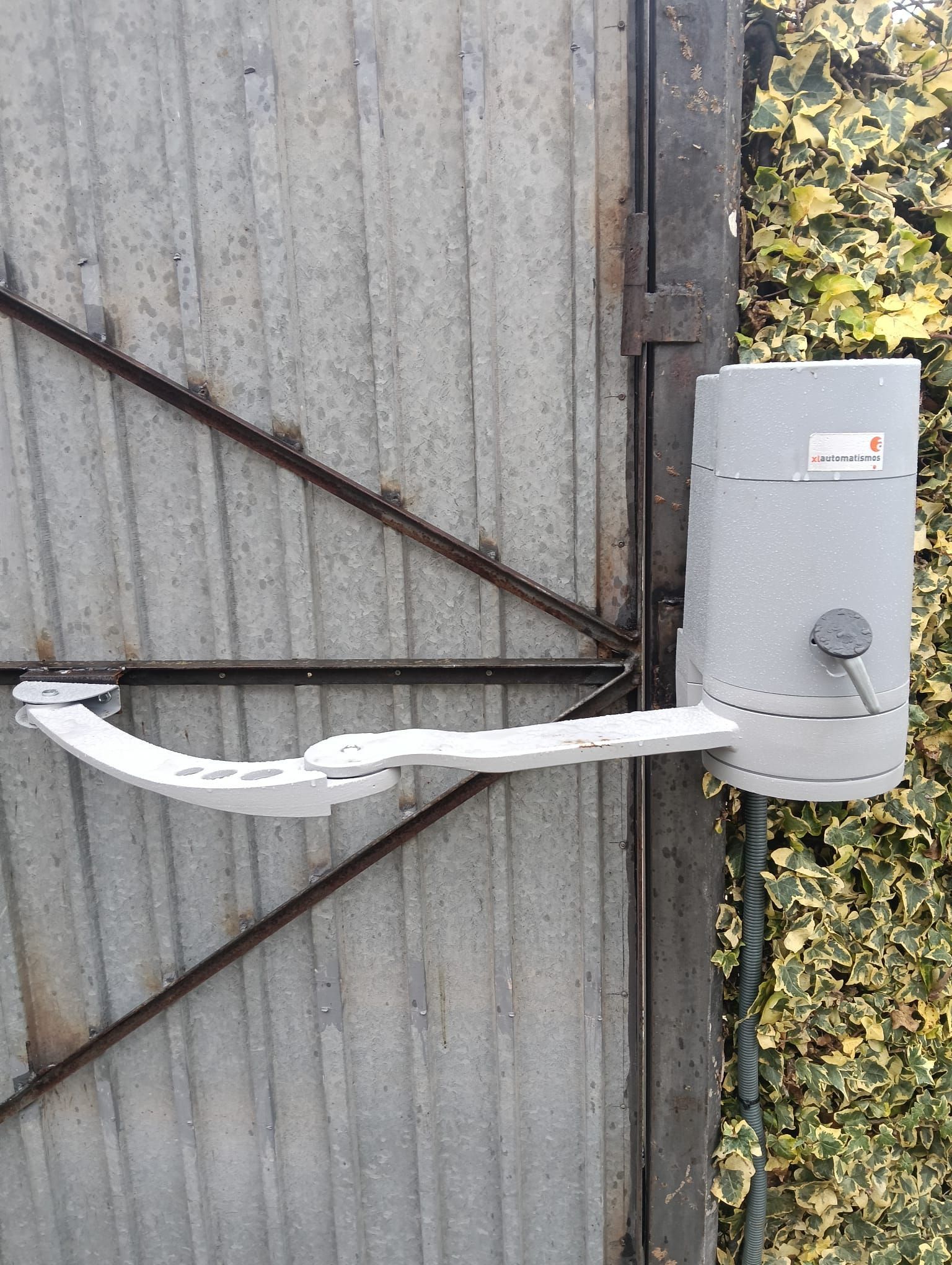 Caja blanca de servicios montada en una puerta metálica ondulada, con un tirador metálico curvo y hiedra a la derecha