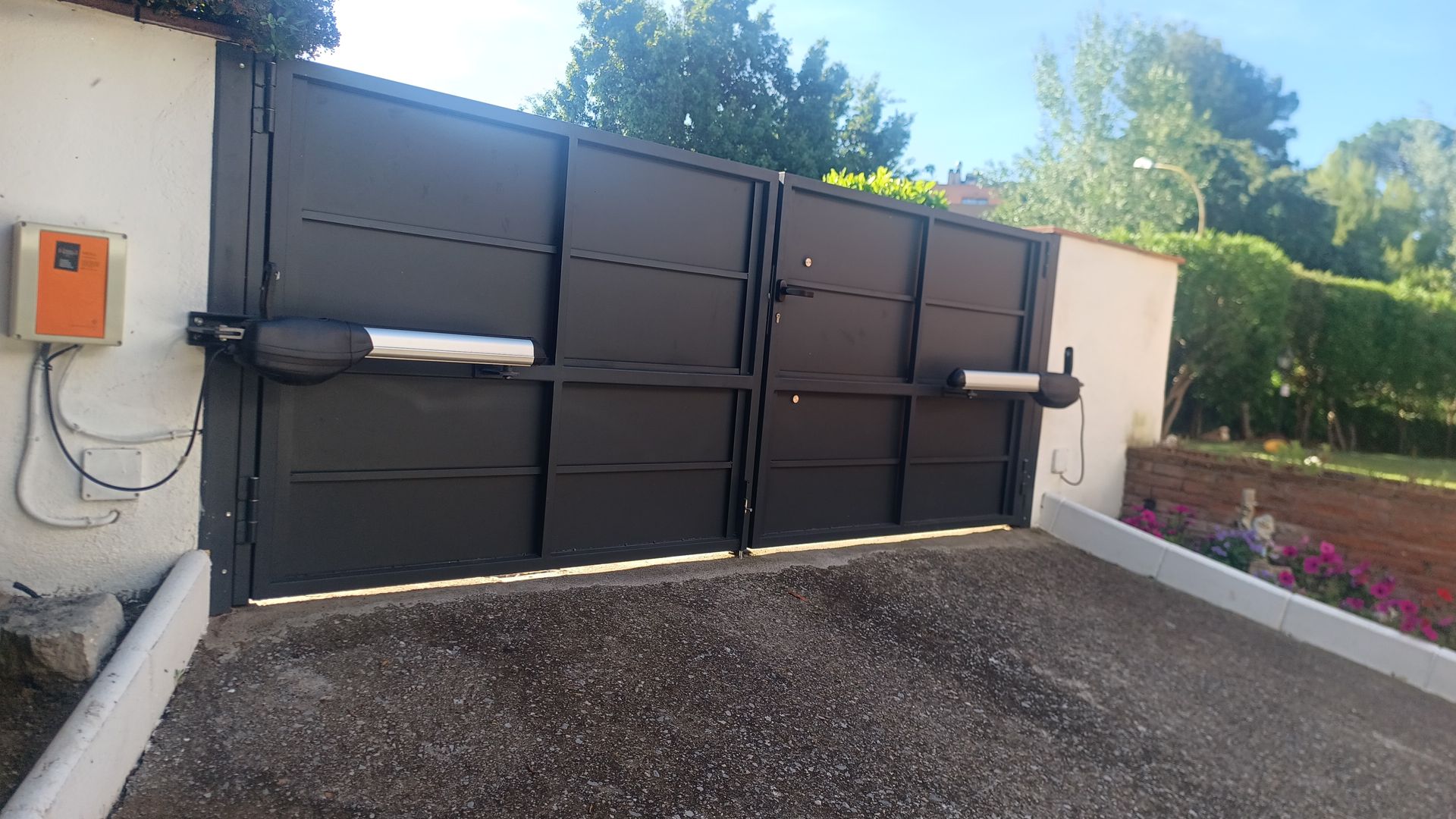 Portón negro cerrado junto a un camino de grava, con una caja de control montada en la pared y un parterre cerca