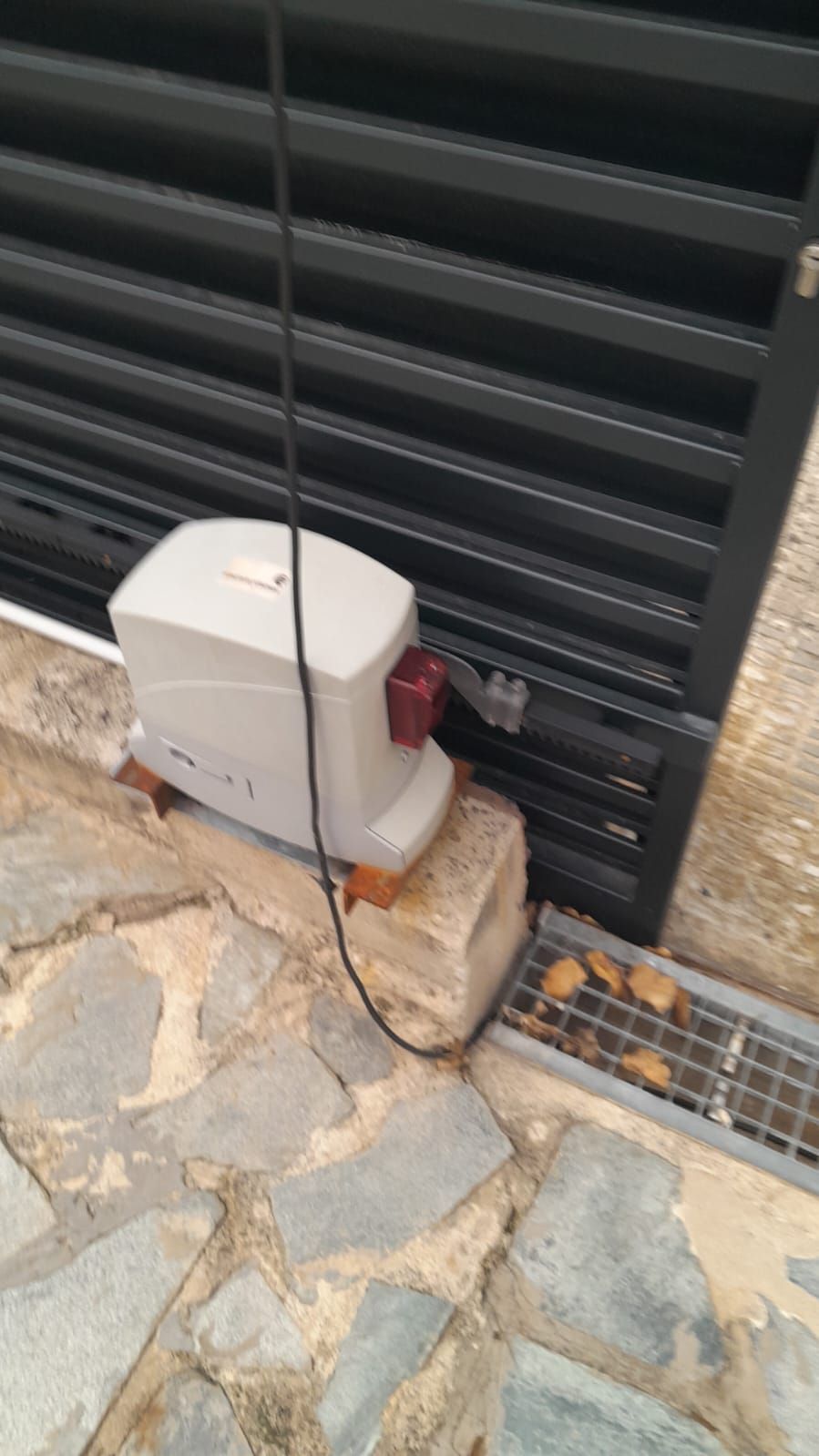 Motor gris de portón junto a un portón metálico negro en una entrada de piedra, con una rejilla de desagüe cerca.