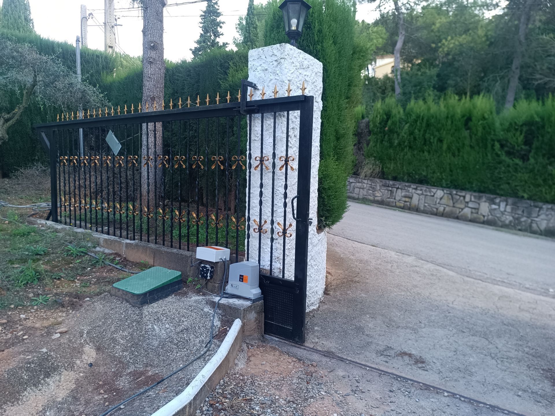 Una verja de metal negro con pilares de piedra da paso a un camino de entrada, junto a un muro de piedra y vegetación.