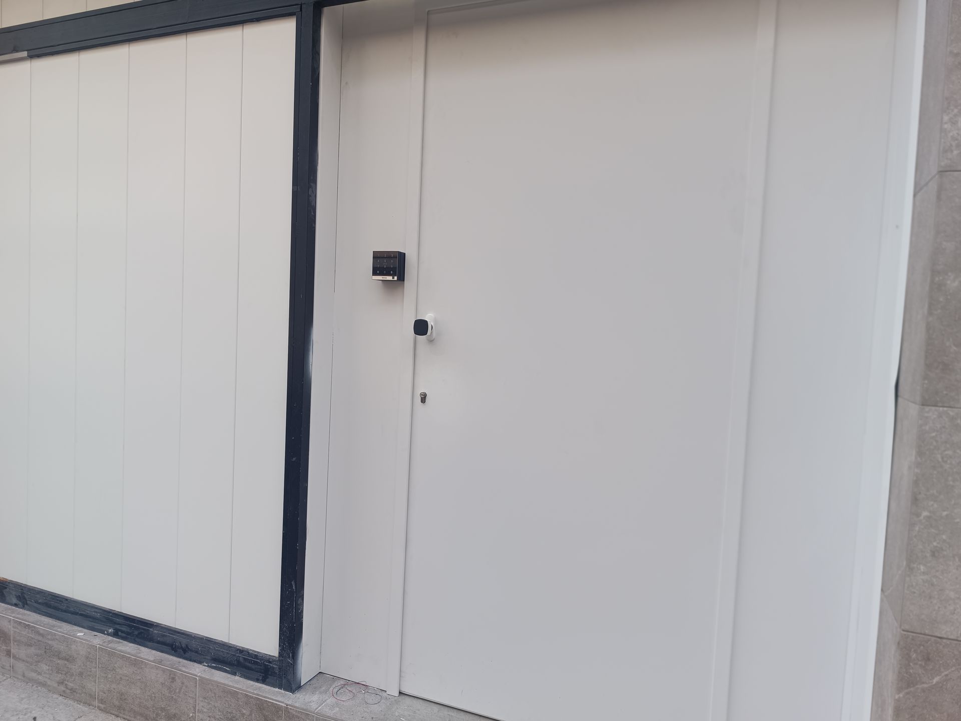 Pared exterior blanca con un pequeño aplique negro y una puerta blanca lisa.