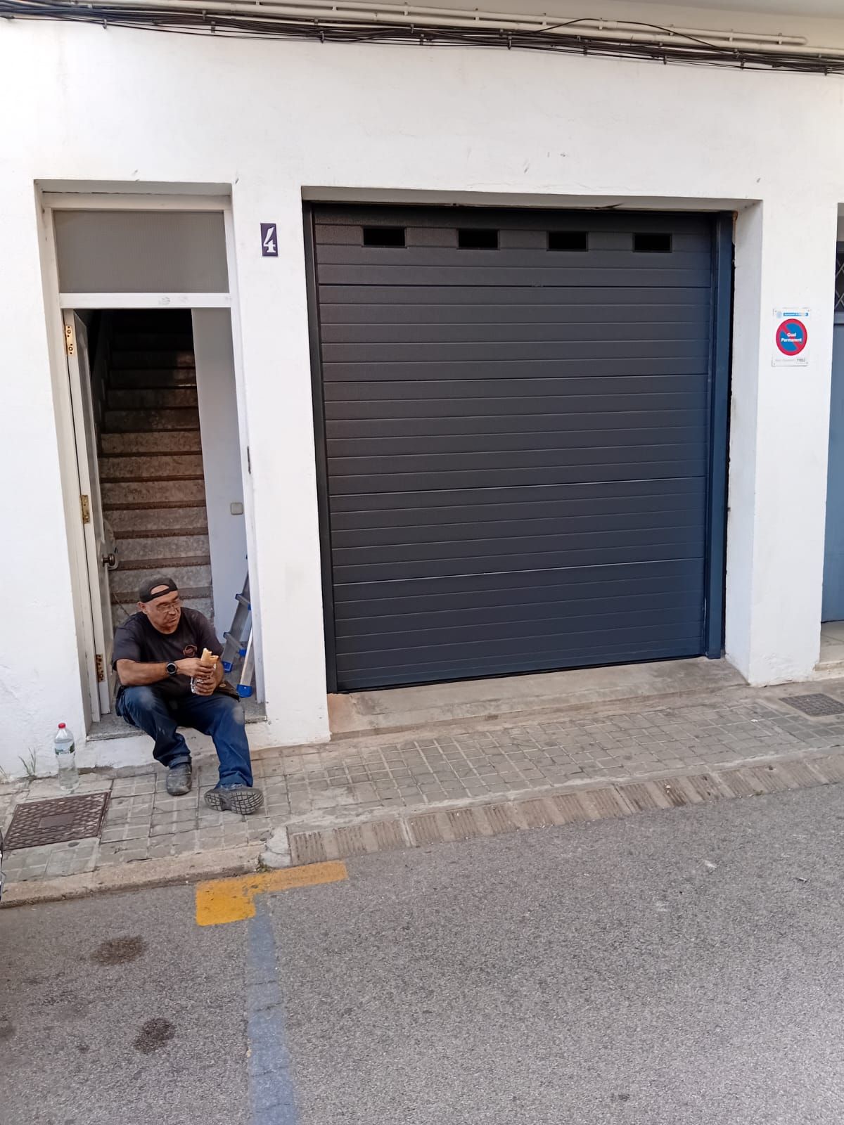 Una persona sentada en el umbral de una puerta, junto a una gran puerta de garaje gris, en una calle tranquila.
