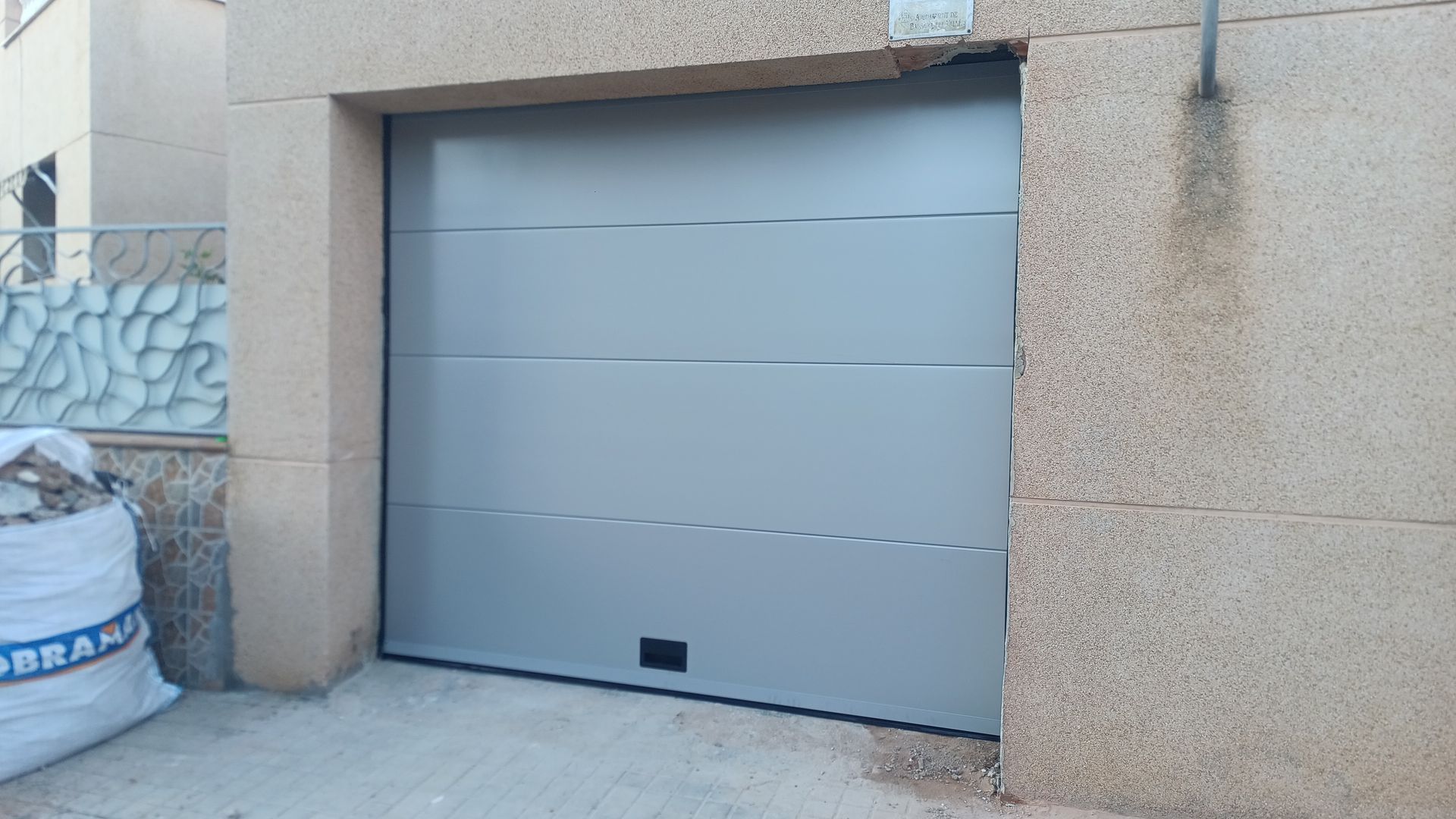 Puerta de garaje gris en una pared de estuco beige, con una pequeña rejilla de ventilación cerca de la parte inferior.