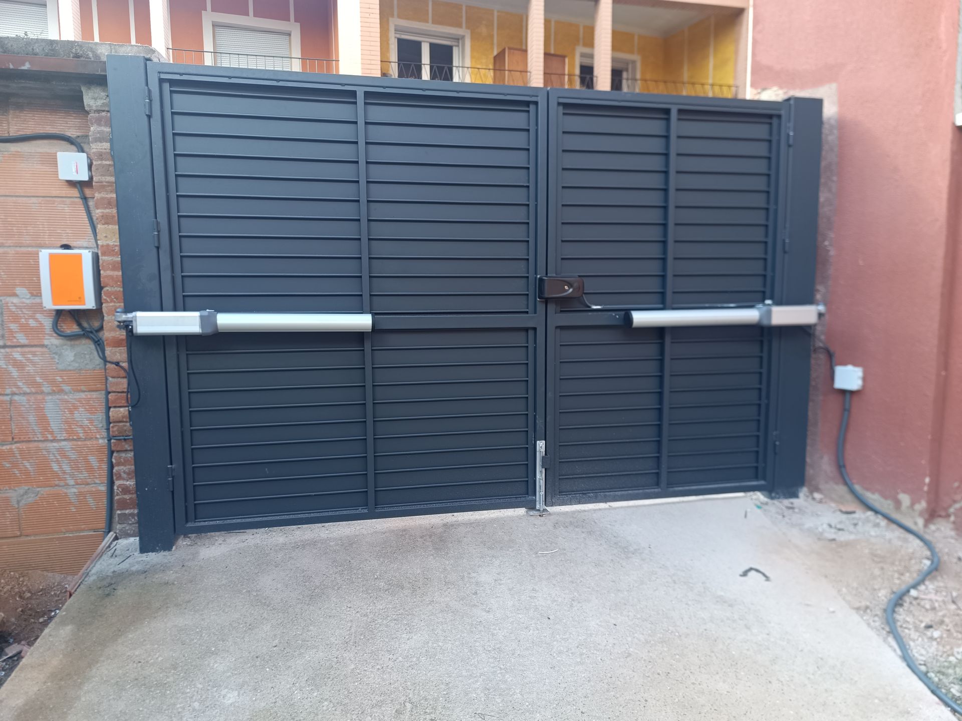 Portón doble de metal negro frente a una casa, con entrada de hormigón y muros laterales