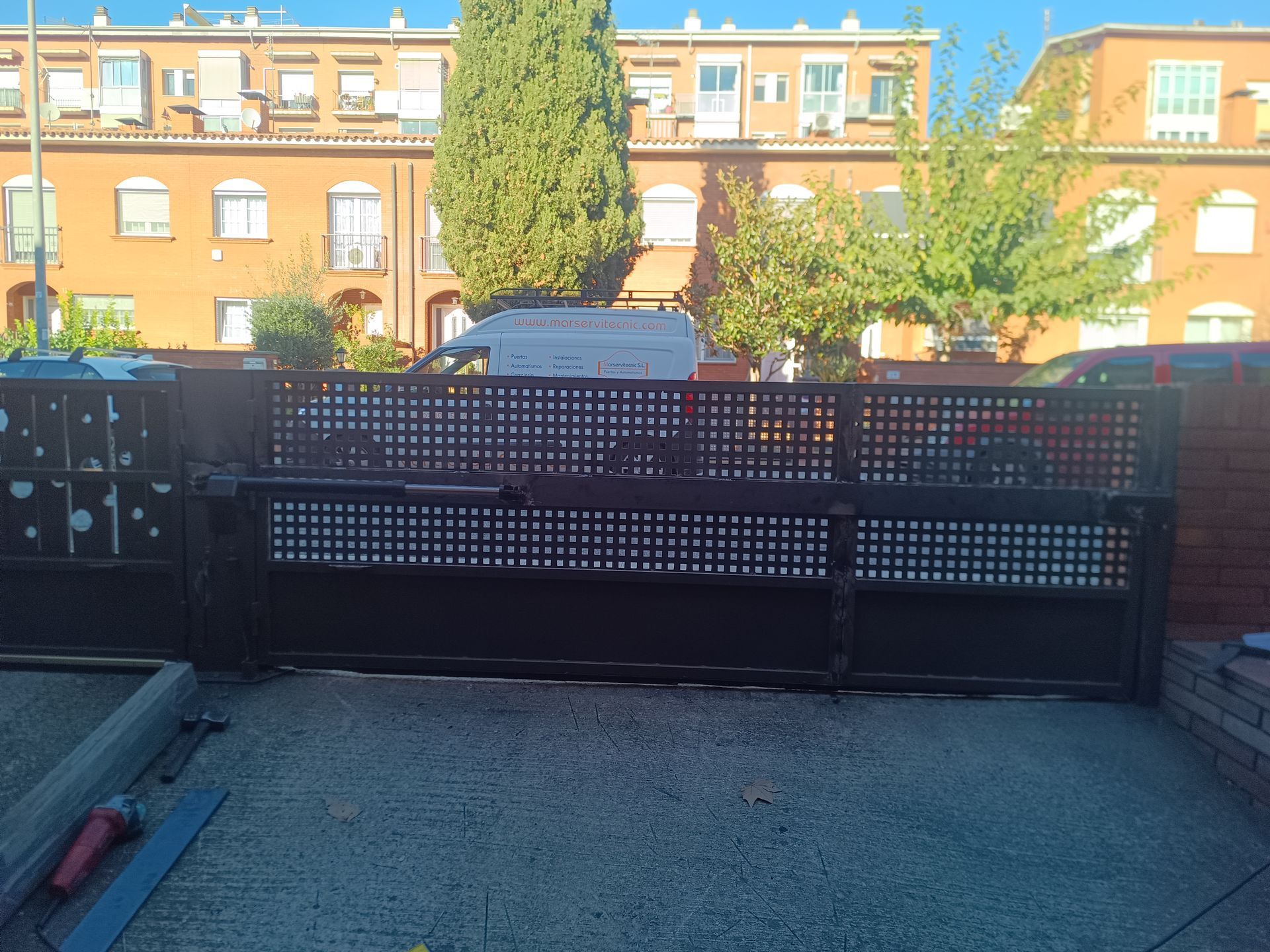 Portón de entrada de metal negro entre muros de hormigón bajo un sol radiante.