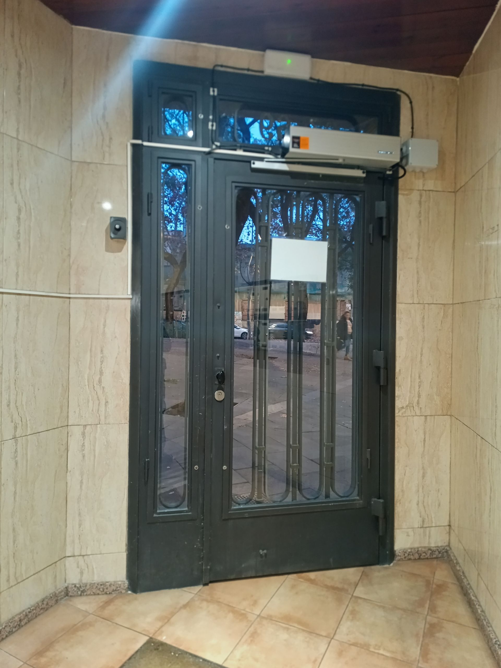 Puerta de entrada de vidrio con barrotes verticales y cierrapuertas superior, en un vestíbulo interior con suelo de baldosas