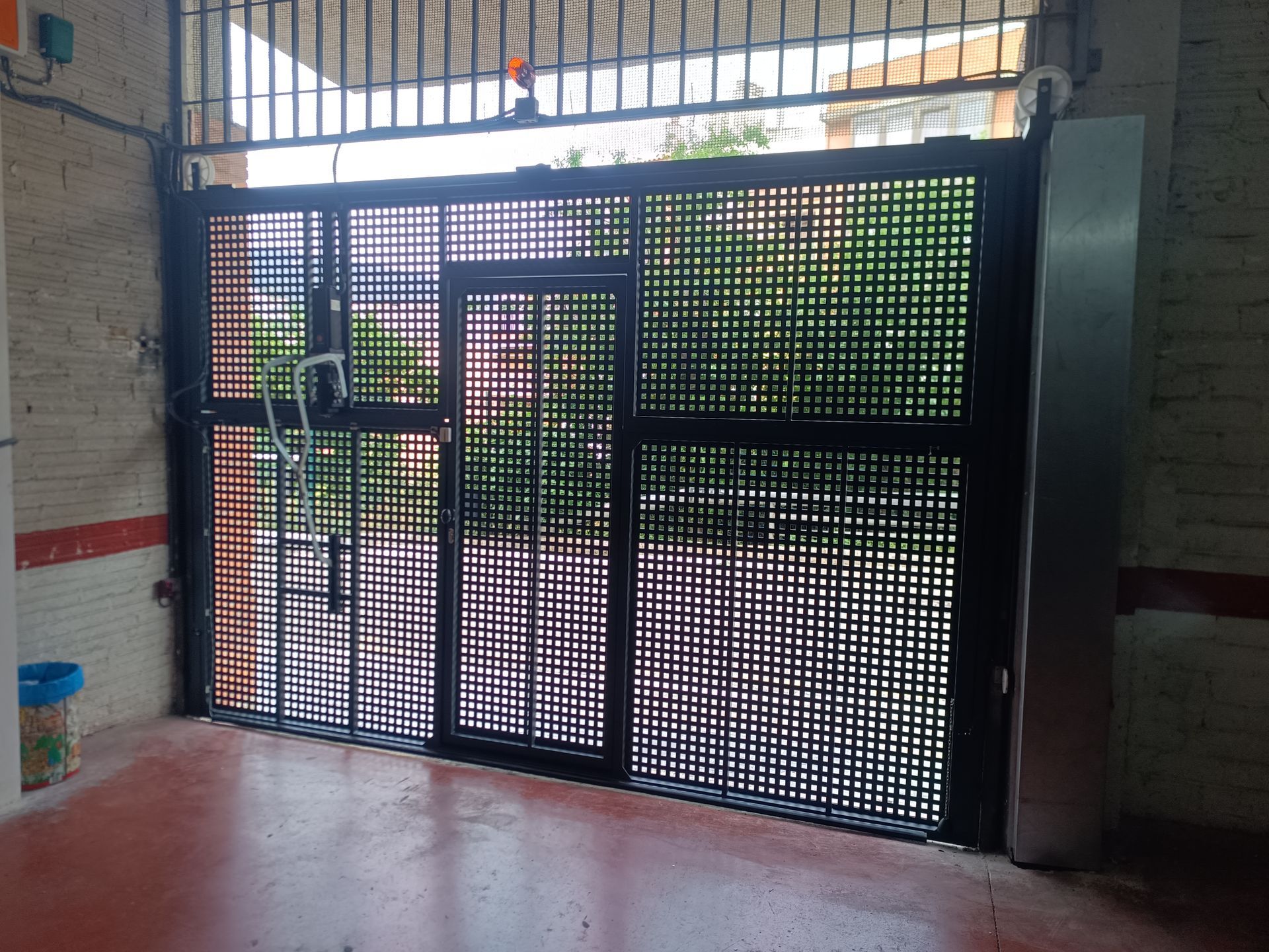 Una puerta de reja negra con una puertecita en la entrada de un edificio, vista desde el interior de un pasillo embaldosado.