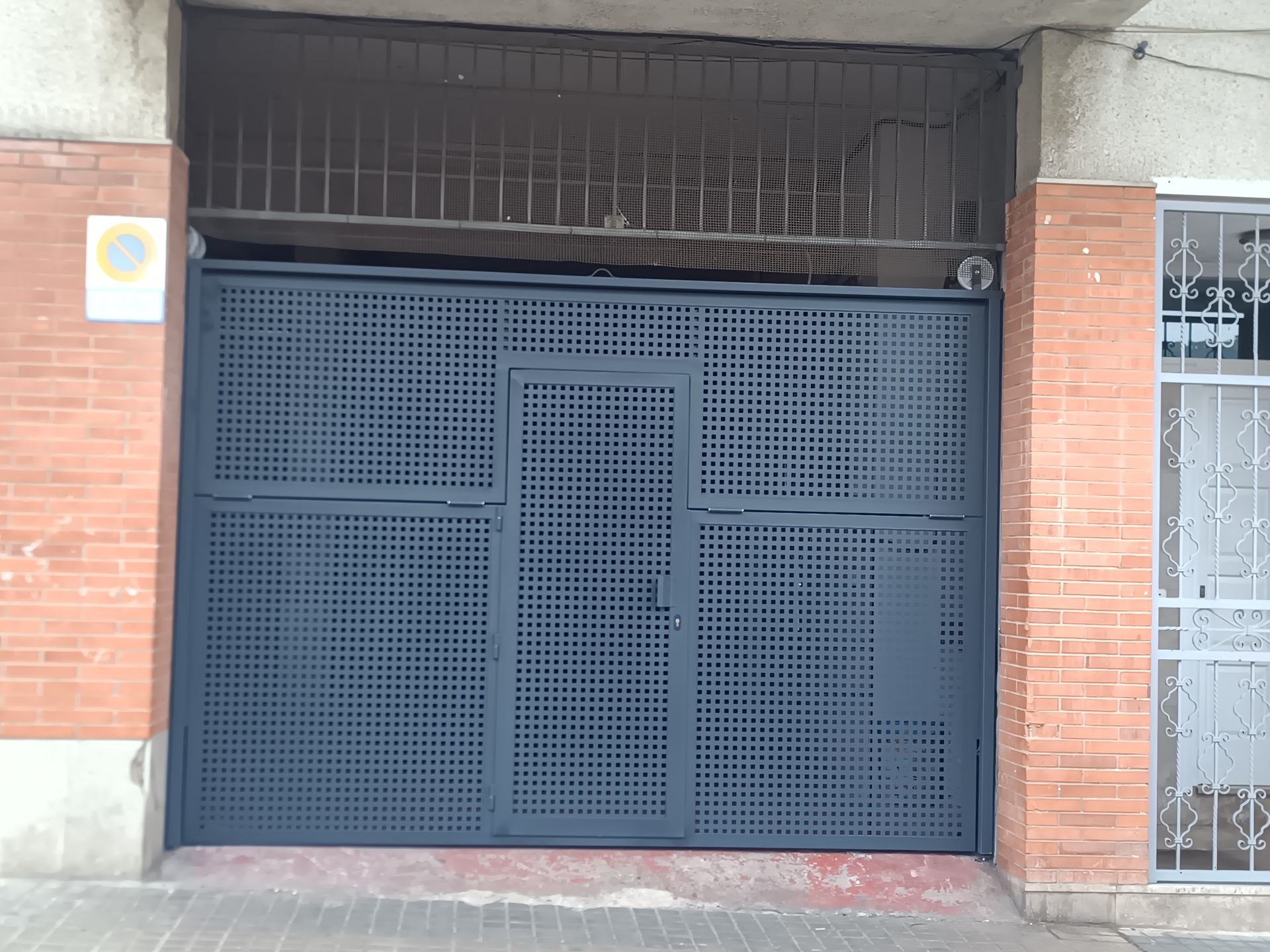 Portón metálico azul cerrado bajo la entrada de un edificio de ladrillo, con una ventana enrejada encima.