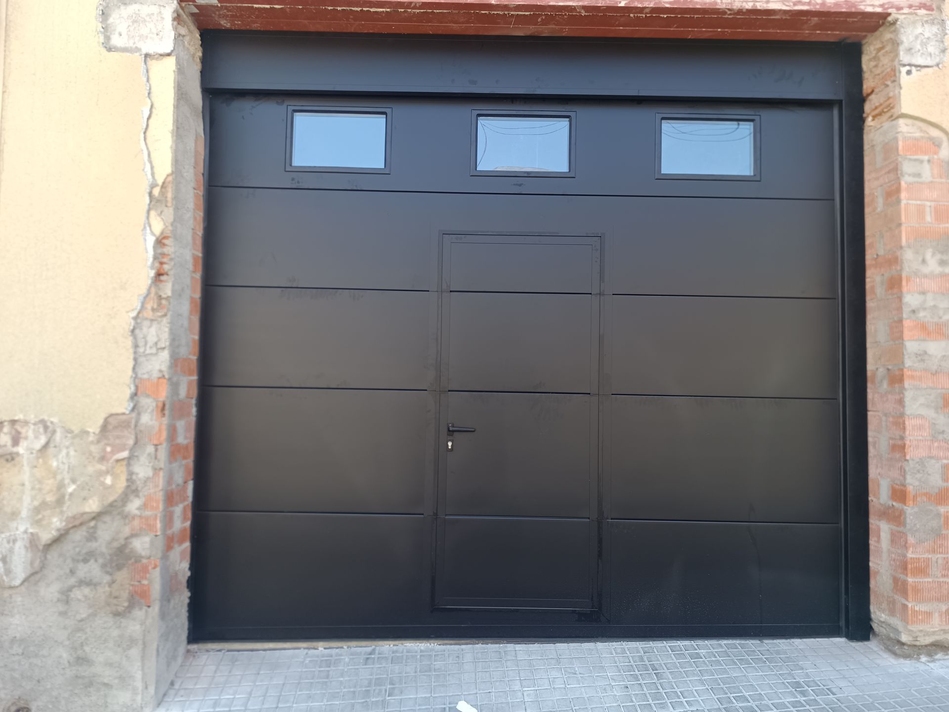 Puerta de garaje negra con tres ventanitas en una pared desgastada de ladrillo y estuco