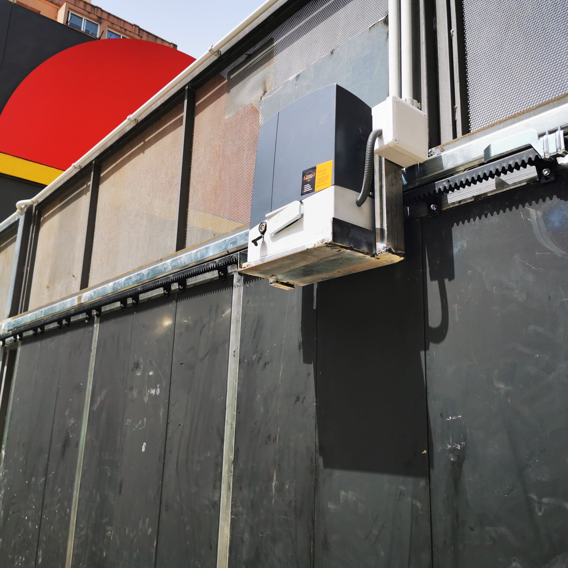 Pared exterior industrial con una caja metálica instalada y paneles de barrera negros bajo un letrero rojo
