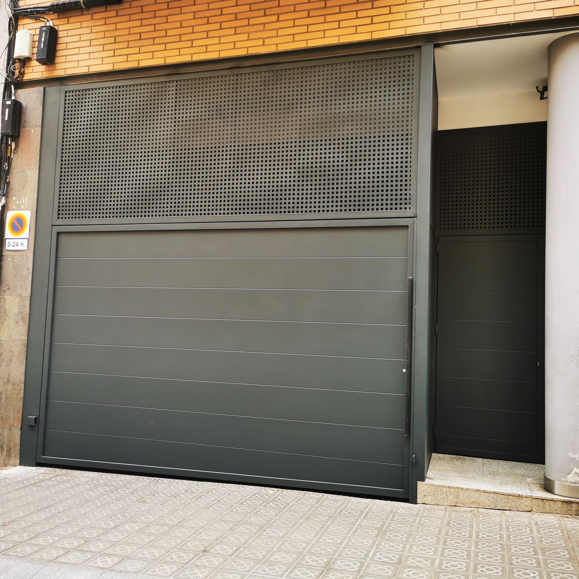 Puerta de garaje metálica cerrada de color gris oscuro en la fachada de un edificio con ladrillo y entrada embaldosada.