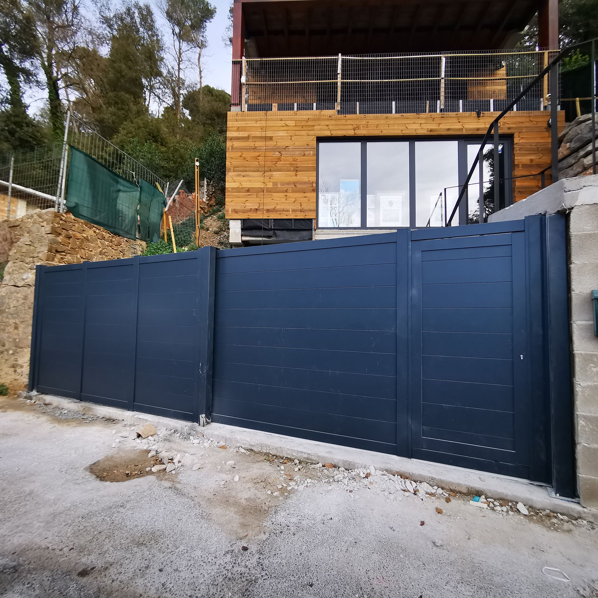 Portón metálico azul oscuro frente a una casa moderna en construcción, con una terraza de madera en la planta superior.