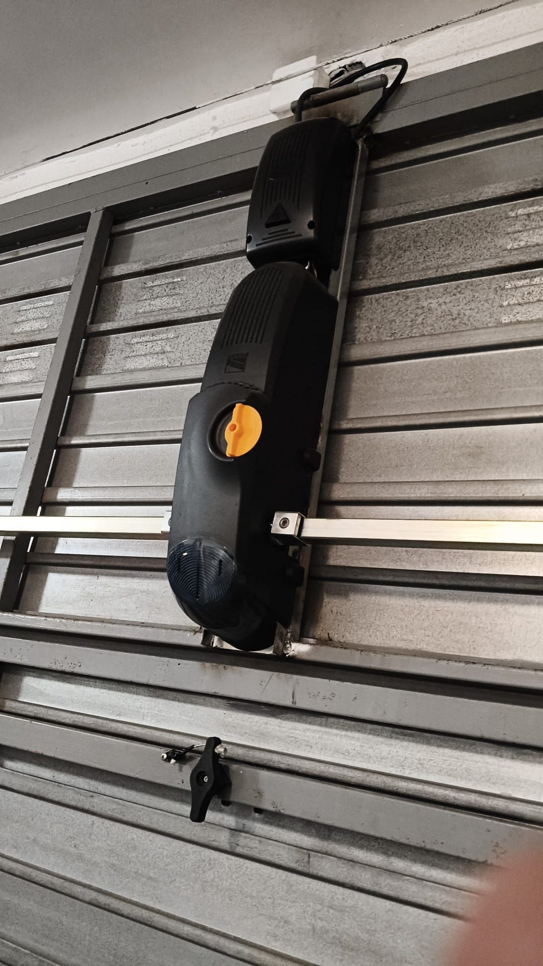 Cámara de seguridad negra instalada en una pared, encima de una persiana metálica de lamas, con una etiqueta naranja.