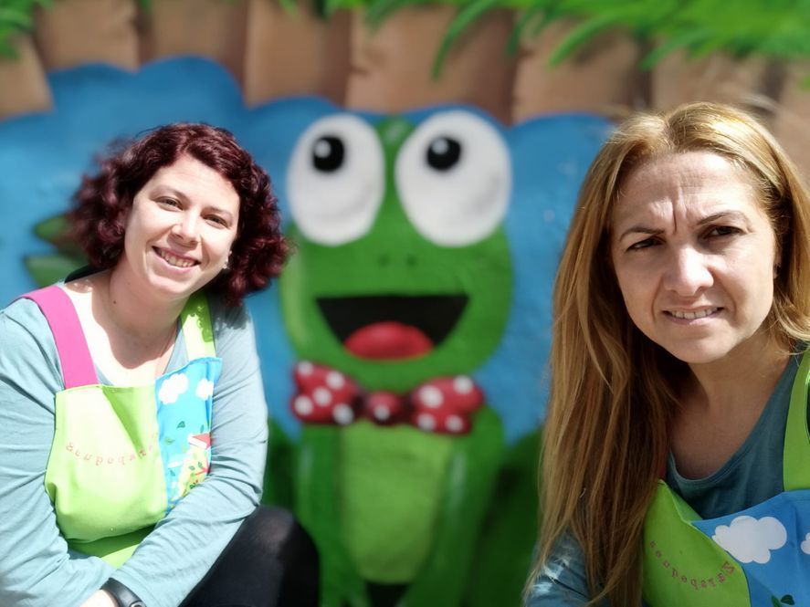 Dos mujeres posan para una foto frente a un mural de ranas.