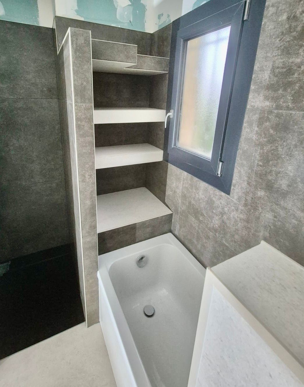 Une salle de bains avec une baignoire blanche, des murs carrelés gris foncé et une étagère intégrée