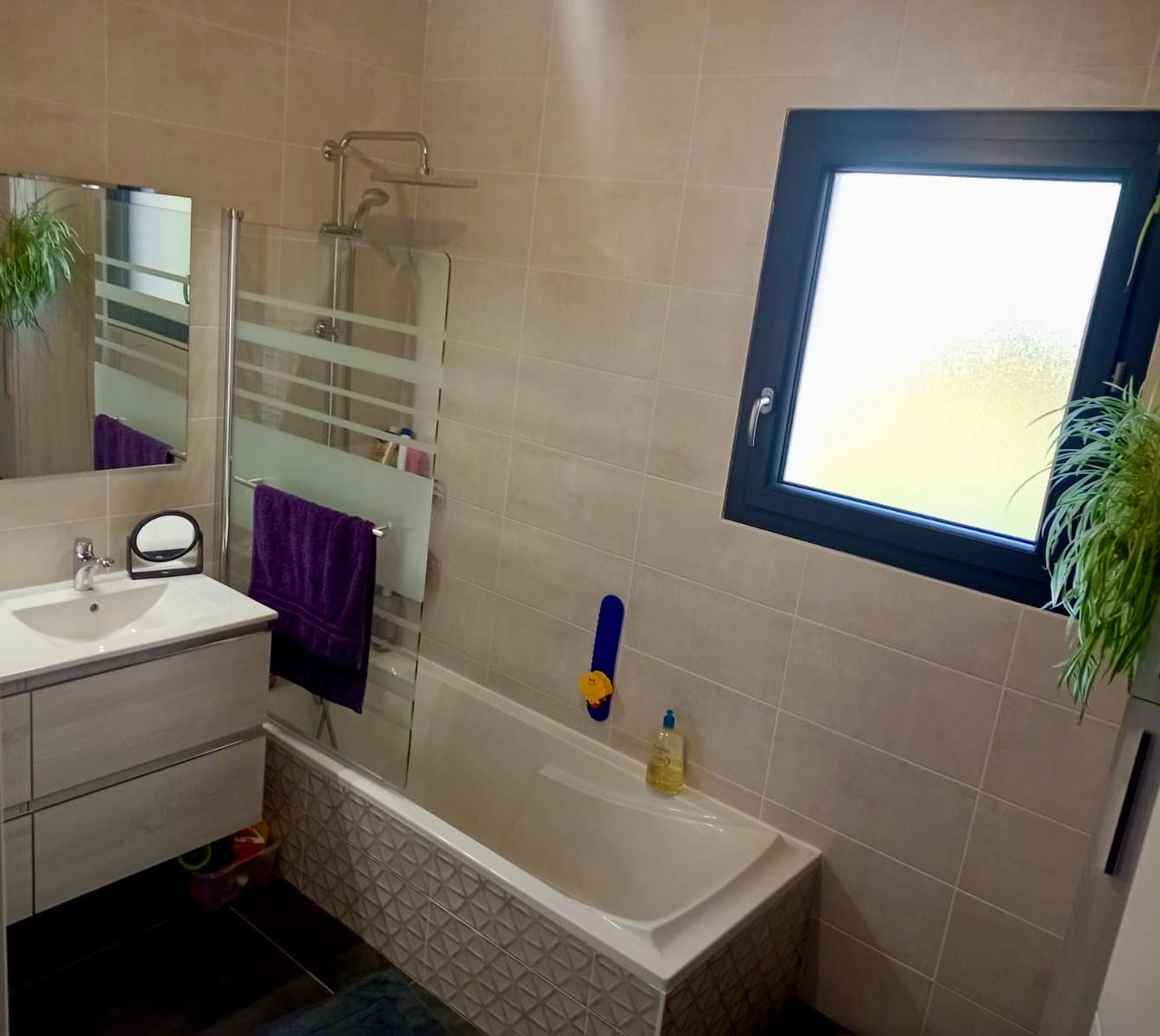 Salle de bains moderne avec meuble-lavabo blanc, baignoire avec paroi de douche en verre, murs carrelés beiges