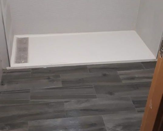 Base de ducha de color blanco instalada en un baño con suelo de baldosas de imitación madera gris.