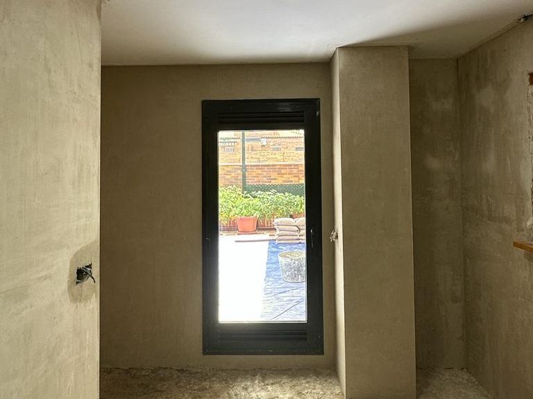 Habitación interior en construcción, pequeña ventana negra, paredes de color claro, vista al jardín exterior.