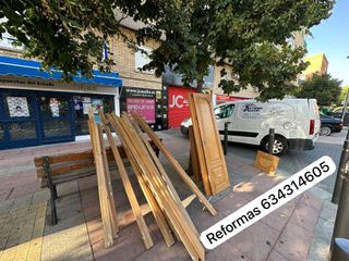 Materiales de construcción de madera apilados en una acera, con un banco, un edificio y una camioneta visibles.