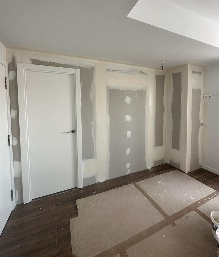 Interior de una habitación en construcción. Paredes de yeso, puerta blanca y suelo de madera oscura.