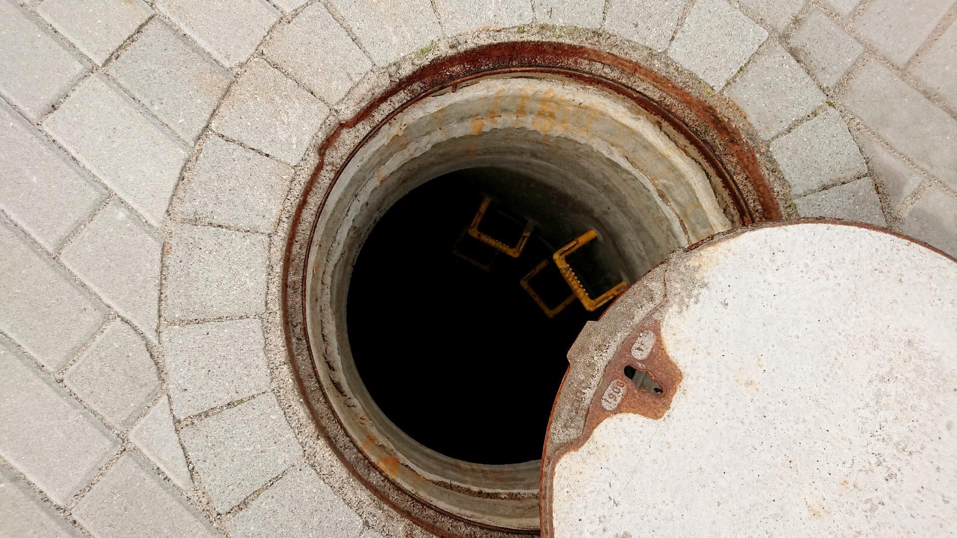 Ein offener Kanaldeckel in einer hellgrauen Pflasterfläche gibt den Blick auf eine dunkle unterirdische Öffnung frei.