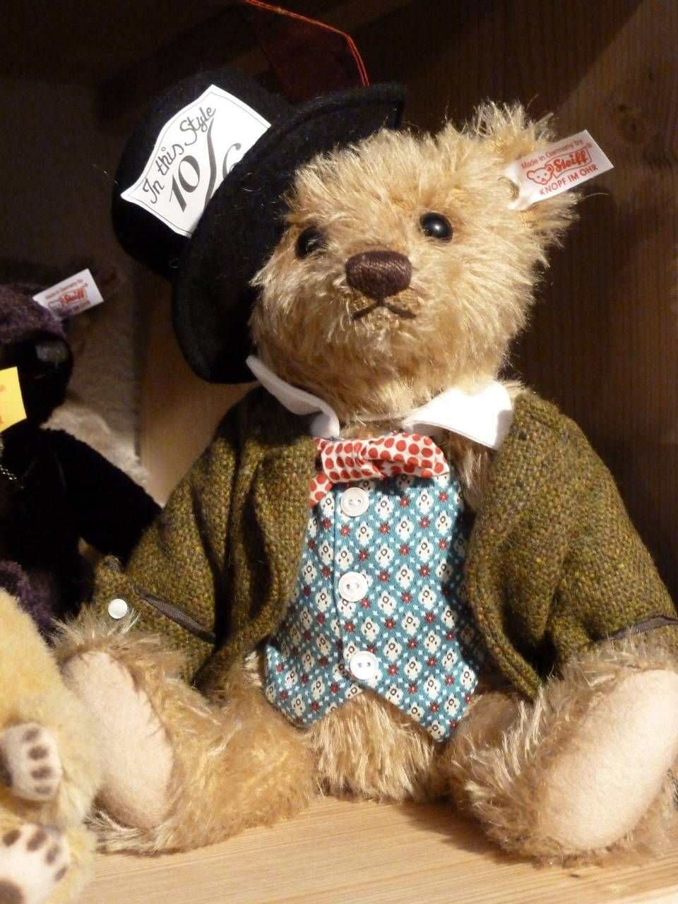 Ours en peluche portant un manteau marron, un gilet bleu et un nœud papillon, coiffé d'un chapeau noir avec une étiquette.