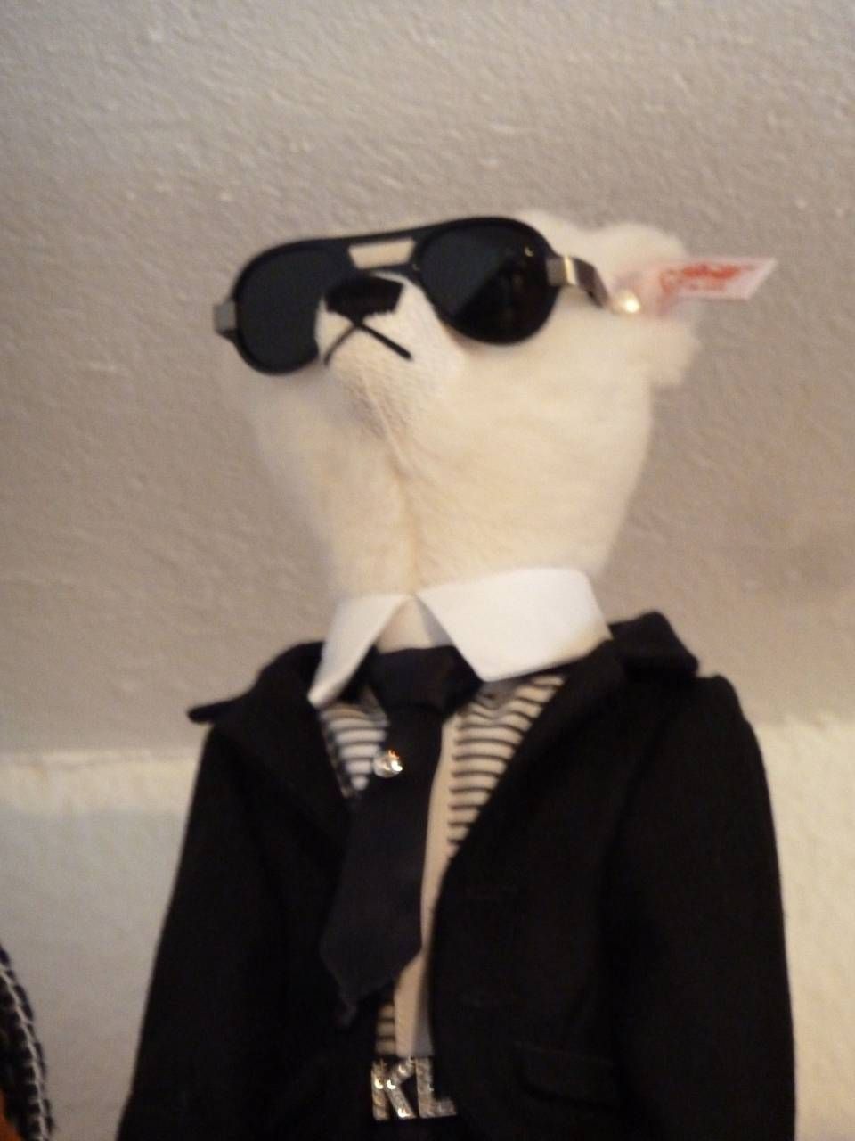 Un alpaga en peluche vêtu d'un costume noir, d'une chemise blanche, d'une cravate rayée et de lunettes de soleil, devant un mur beige.