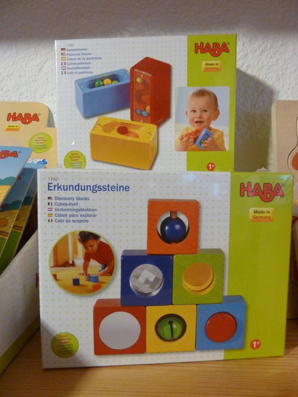Des boîtes à jouets HABA colorées sont empilées sur une table, représentant un bébé et des blocs de formes en bois à trier.
