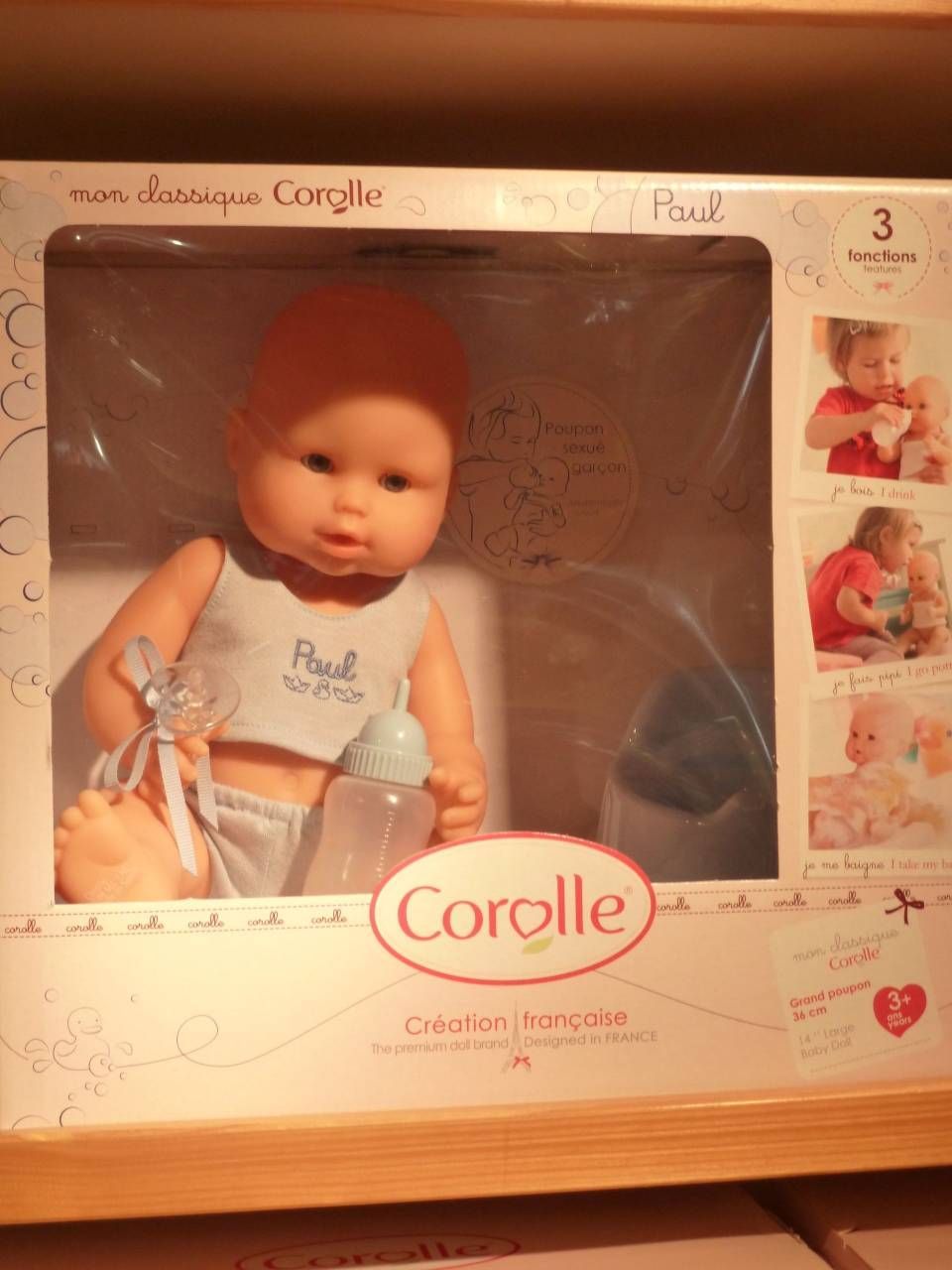 Poupée Corolle dans une boîte cadeau rose et crème, vêtue d'une tenue blanche avec chapeau et biberon assortis.