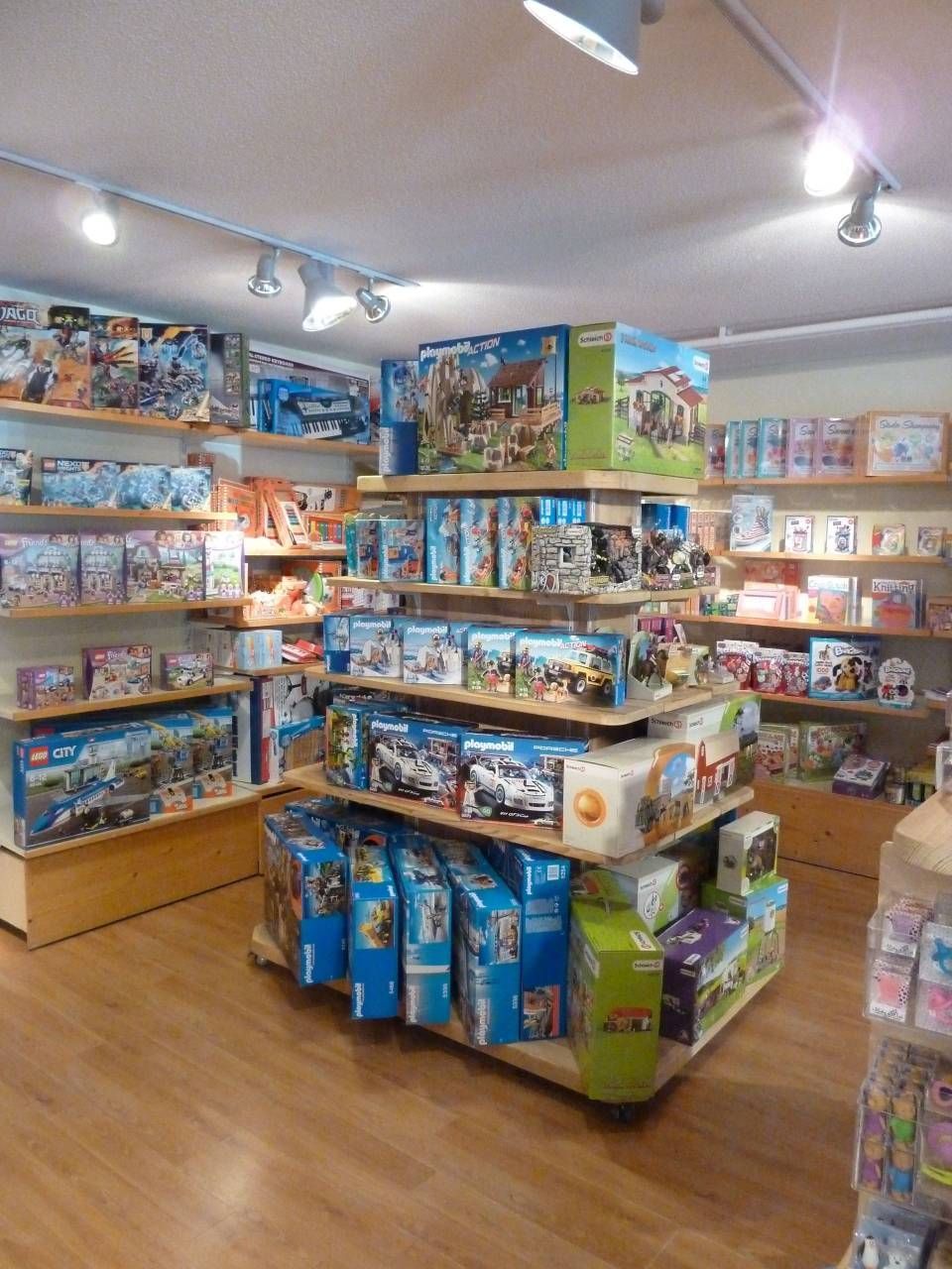 Allée d'un magasin de jouets bien éclairée, avec des étagères remplies de jouets et de jeux colorés en boîte, posées sur un sol en bois.