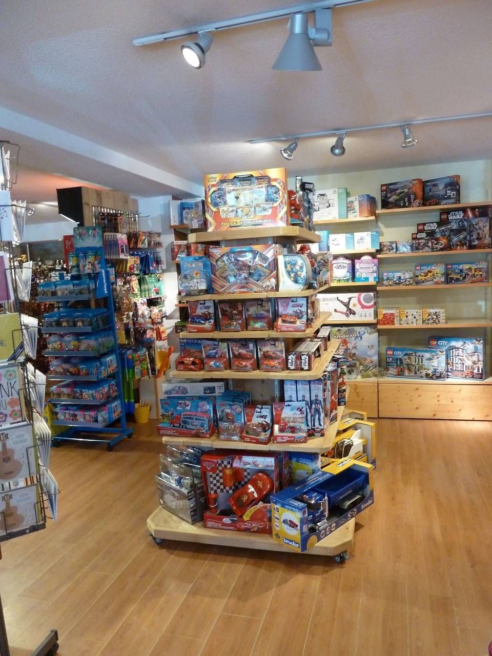 Allée d'un magasin de jouets avec des jeux de société et des jouets colorés sur des étagères en bois, sous des projecteurs puissants.
