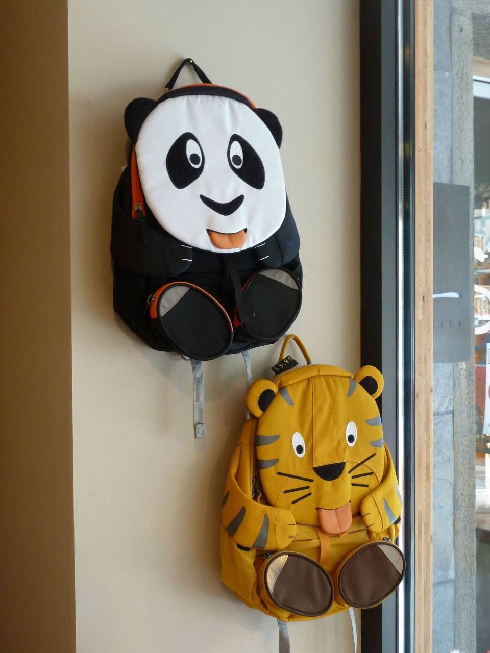 Deux sacs à dos en forme d'animaux, un panda et un tigre, sont accrochés à des crochets muraux près d'une fenêtre.