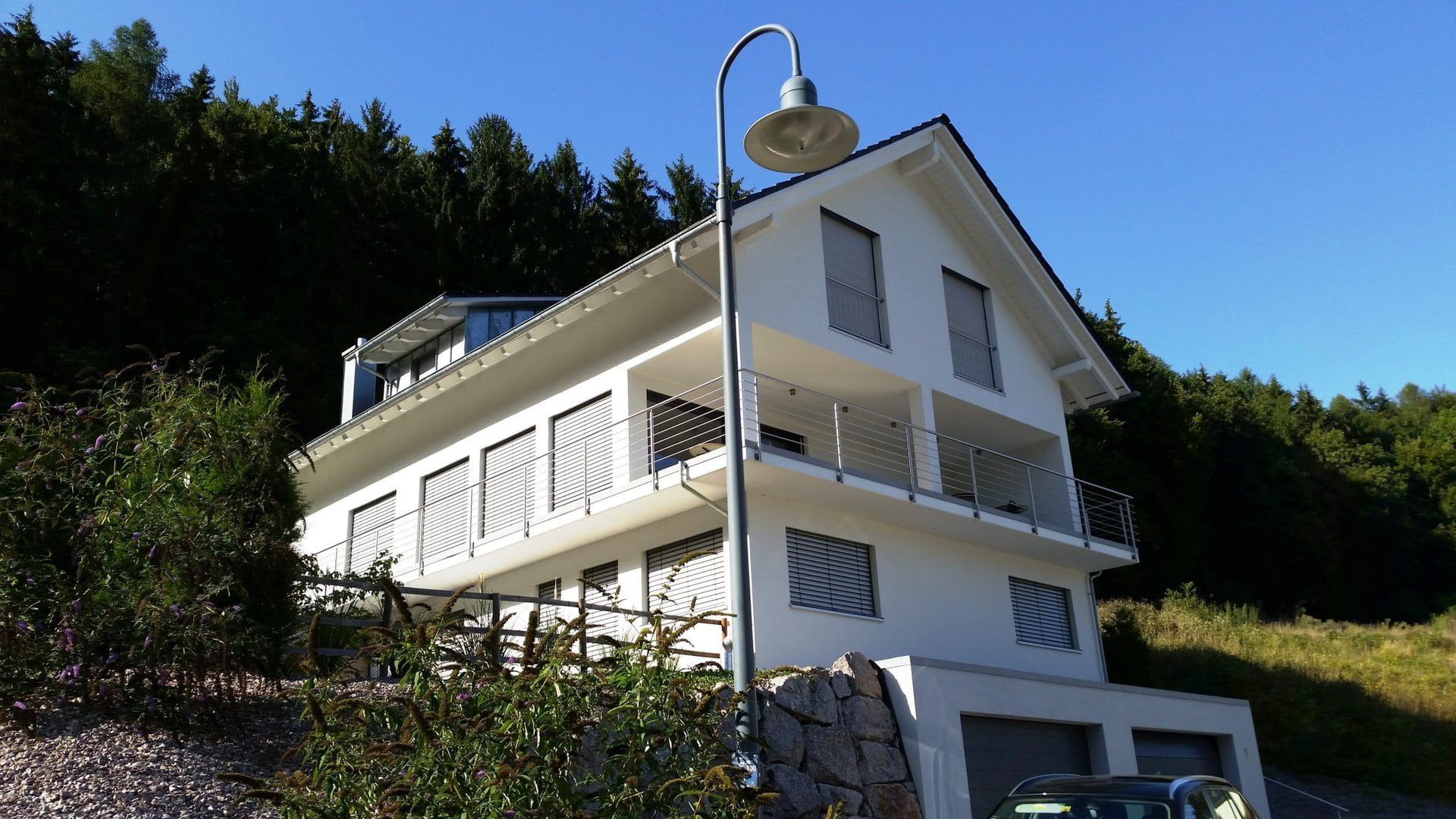 Weißes Haus mit Balkonen, Garage und Waldkulisse unter blauem Himmel.