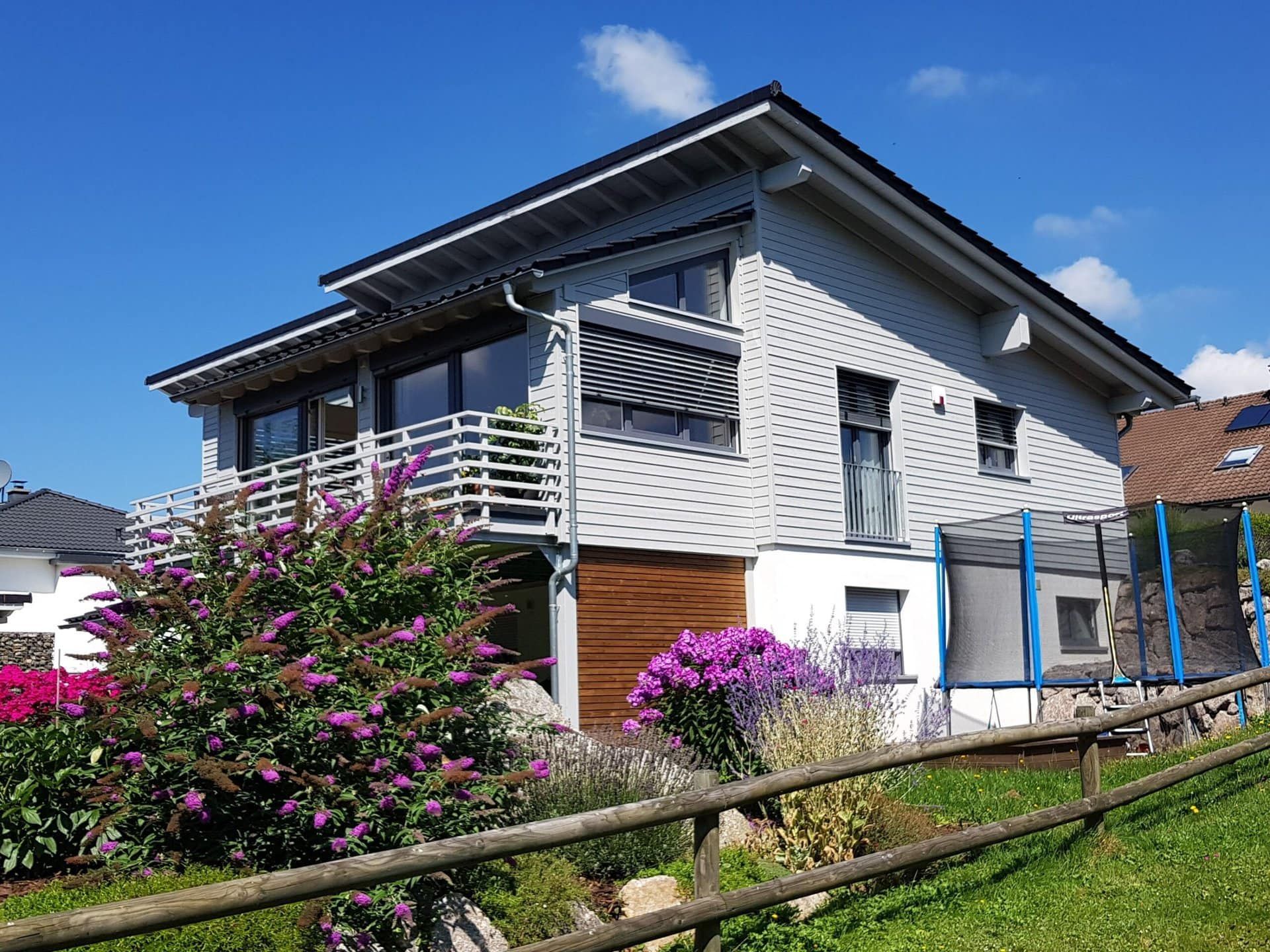 Modernes zweistöckiges Haus mit grauer Fassade, Balkon und Gartengestaltung vor blauem Himmel.
