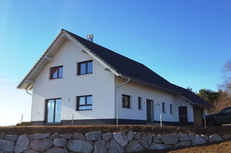 Weißes Haus mit schwarzem Dach und Fenstern vor blauem Himmel, erbaut auf einer steinernen Stützmauer.