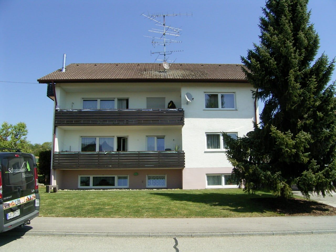 Zweistöckiges Mehrfamilienhaus mit Balkonen, braunem Dach und einem großen immergrünen Baum. Links parkt ein Lieferwagen.