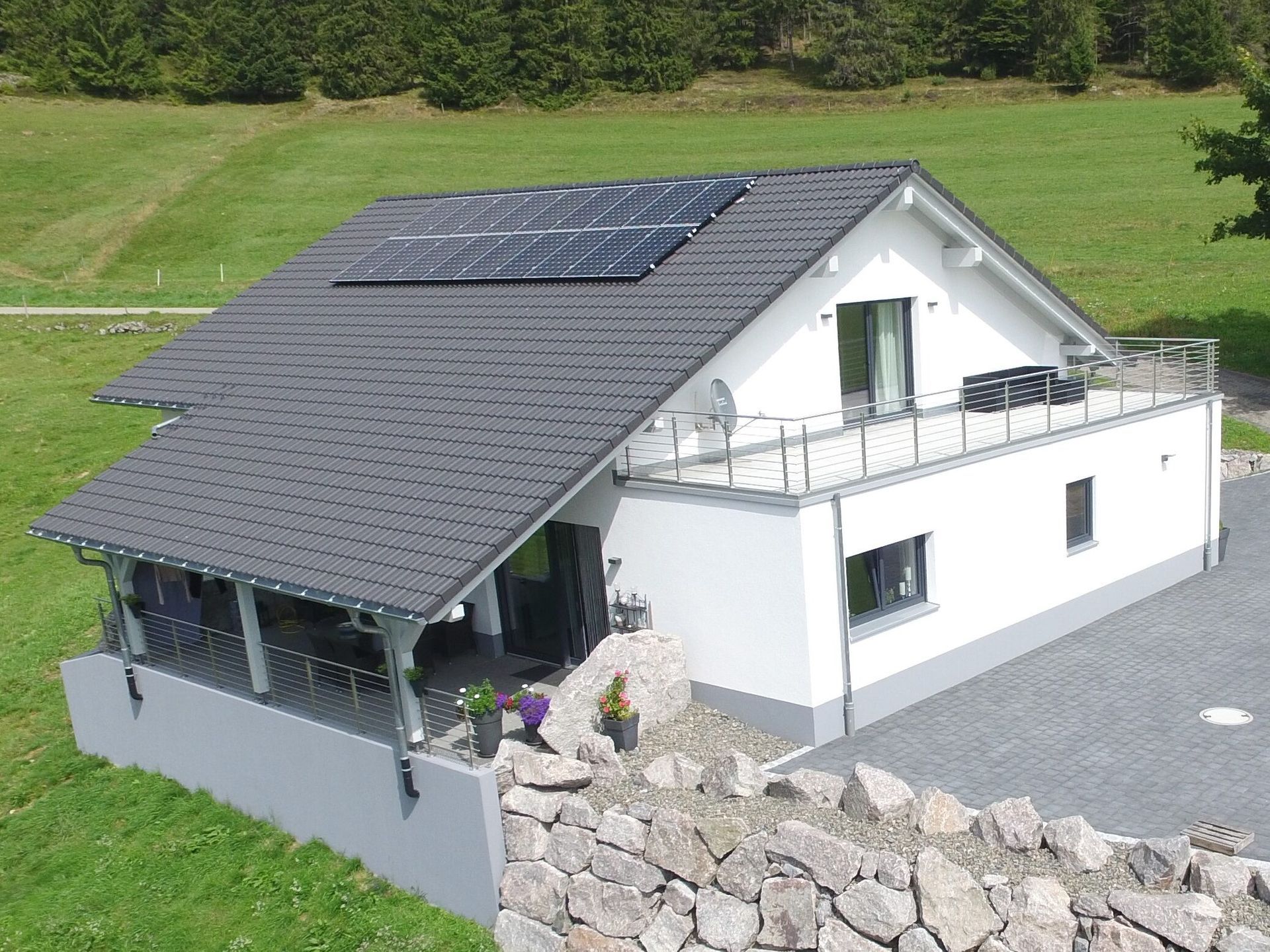 Weißes Haus mit Solarpaneelen auf dunkelgrauem Dach, Balkon und Steinmauer in grasbewachsener Umgebung.