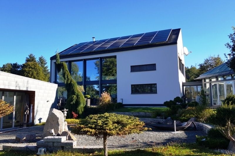 Modernes weißes Haus mit Solarpaneelen, großen Fenstern und einem angelegten Garten an einem sonnigen Tag.