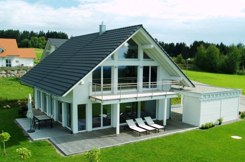 Modernes weißes Haus mit dunkelgrauem Dach, großen Fenstern, Balkon und grünem Rasen.