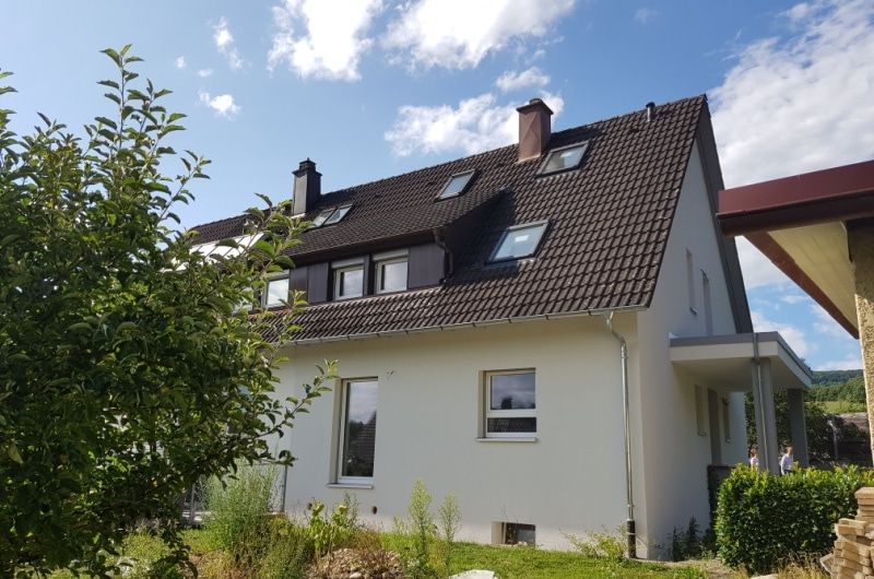 Weißes Haus mit dunklem Dach und mehreren Dachfenstern, sonniger Tag mit blauem Himmel.