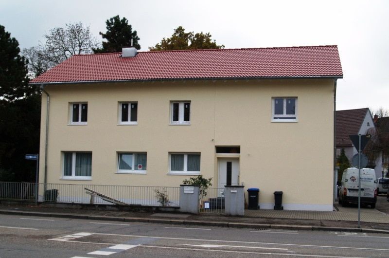 Zweistöckiges Gebäude mit rotem Ziegeldach, beiger Fassade, weiß gerahmten Fenstern und Blick auf die Straße.