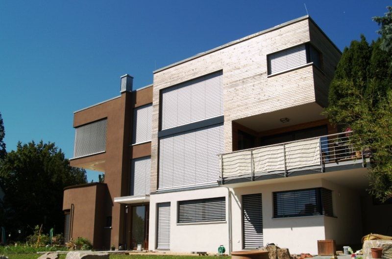 Modernes zweistöckiges Haus mit hellbrauner Holzverkleidung, weißem Stuck und horizontalen Jalousien an den Fenstern unter blauem Himmel.