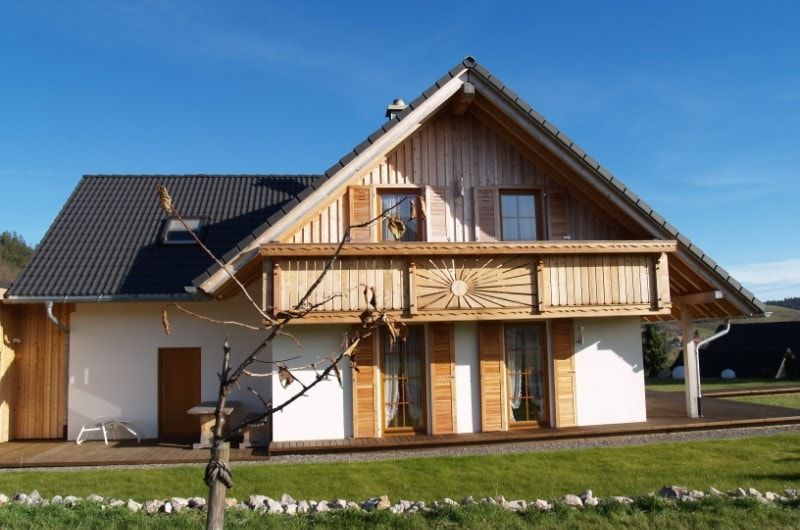 Häuschen mit Holzbalkon und Fensterläden, weißen Wänden und schwarzem Dach vor klarem, blauem Himmel.